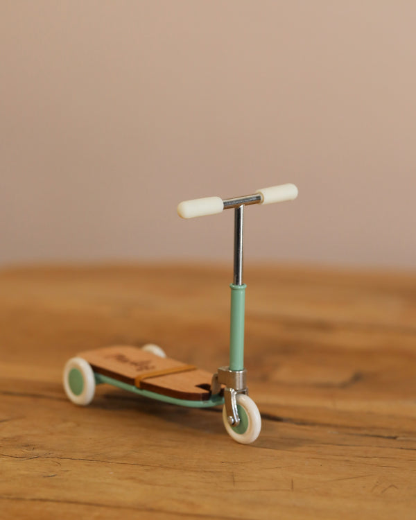 A Mint Maileg Mouse Size Scooter / Kickboard rests on a wooden surface, displaying subtle wooden accents. It features white wheels and a simple handlebar. The neutral background softly blends with the table's tone.