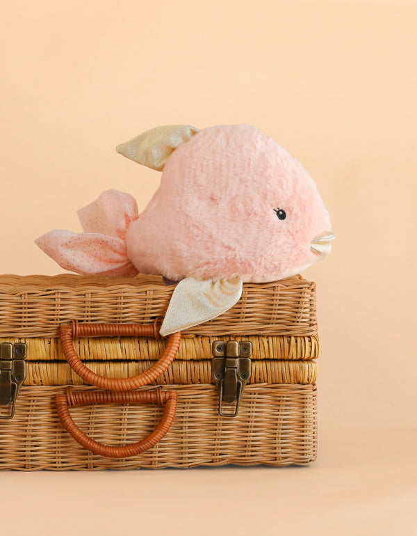 Mon Ami - Crystal the Fish Plush Toy, a soft pink stuffed animal with shiny fins, sits atop a closed wicker suitcase against a beige background. This tropical fish plush makes an adorable gift for kids.