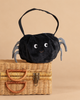 The Mon Ami - Spider Halloween Bag, a small black plush bag with googly eyes and striped legs, sits atop a woven suitcase against a beige background.