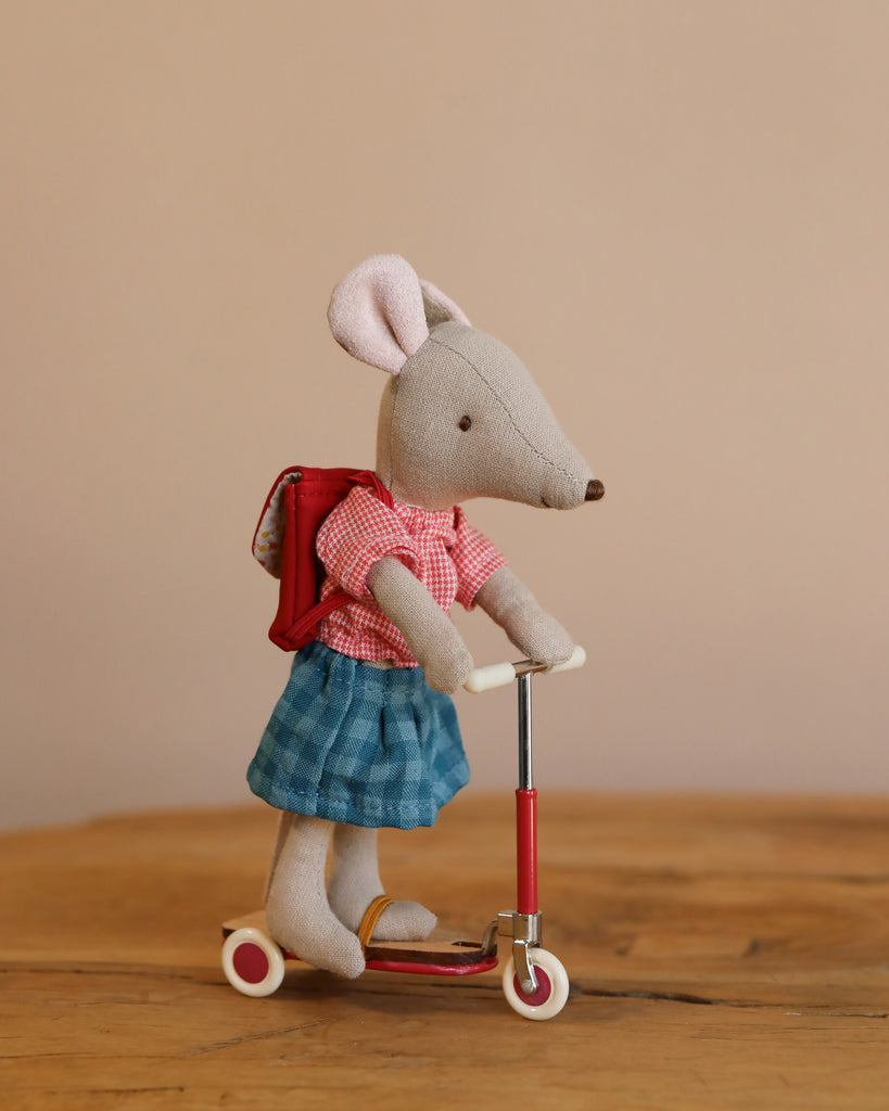 A Maileg Mouse wearing a red backpack, red checkered shirt, and blue skirt stands on a Maileg Mouse Size Scooter / Kickboard - Red, featuring wooden details. The background includes a plain beige wall and wooden floor.