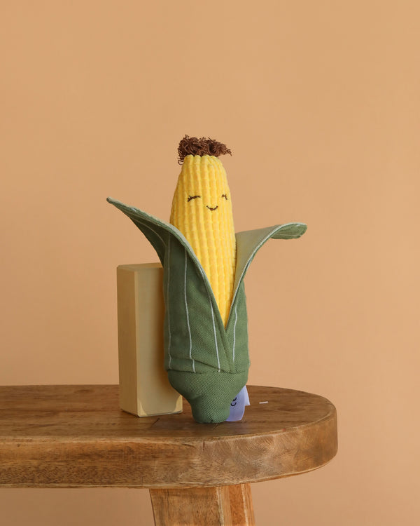 The Cornelius Cob Plush Toy, shaped like a smiling ear of corn with a partially open green husk, sits on a wooden table against a beige background—making it an adorable and cuddly gift for kids.