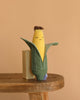 The Cornelius Cob Plush Toy, shaped like a smiling ear of corn with a partially open green husk, sits on a wooden table against a beige background—making it an adorable and cuddly gift for kids.