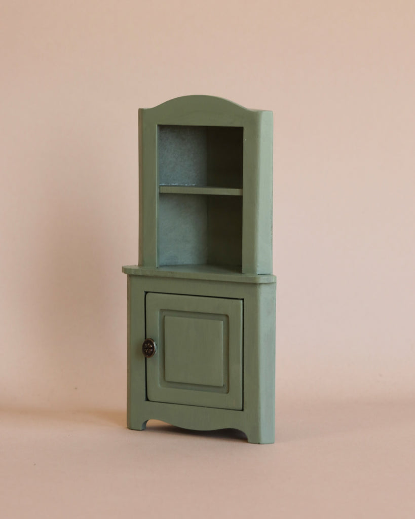 A small green wooden cabinet with one lower door and an open upper shelf displays the Maileg Big Sister Afternoon Tea Set, set against a plain beige background.