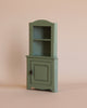 The Maileg Corner Cabinet, Mouse - Light Green is a small wooden piece with an upper open shelf and a lower section featuring a door. Its simple vintage finish adds charm to any home decor. Set against a plain beige background, this cabinet is perfect for cozy spaces needing extra storage.