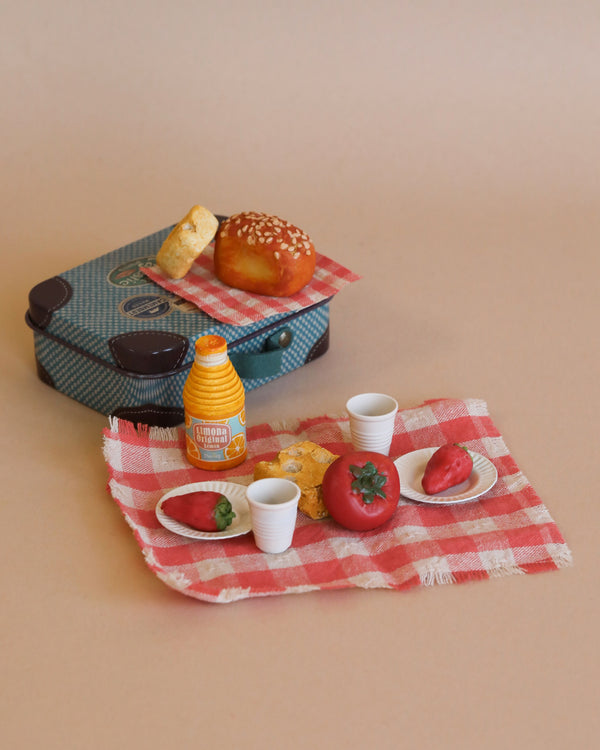 A miniature picnic setup includes tiny food items on a red and white checkered cloth. There are plates with strawberries, coffee cups, a croissant, a tomato, and an orange juice bottle. A small blue suitcase with a magnetic lock houses a bread roll and another croissant, making it the perfect Maileg Picnic Set, Mouse Size dollhouse accessory.