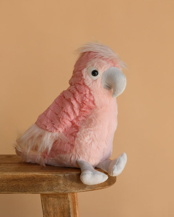 The Lulu Cockatoo Bird Plush Toy, featuring pink and white feathers and a large gray beak, sits on a wooden surface against a plain beige background—a delightful gift for any bird plush lover.
