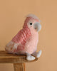 The Lulu Cockatoo Bird Plush Toy, featuring pink and white feathers and a large gray beak, sits on a wooden surface against a plain beige background—a delightful gift for any bird plush lover.
