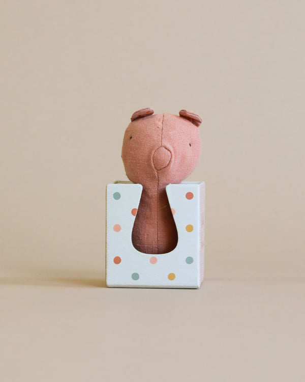 A small, pink Maileg Lullaby Friend Rattles, Pig with two ears sits facing away inside a white wooden gift box with polka dots of various colors. The background is a neutral beige, complementing the soft fabrics perfectly.