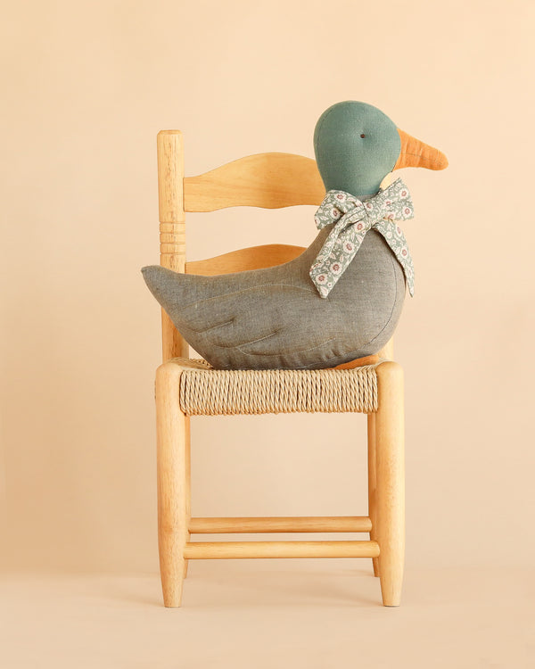 The Maileg Duck with Bow - Grey, featuring a green head, orange beak, and floral bow, is perched on a light wooden chair against a beige background—making it a charming Christmas duck decoration that brings an authentic festive atmosphere to your seasonal animal collection.