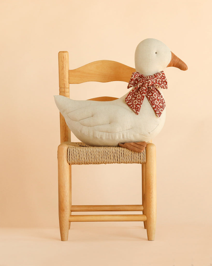 The Maileg Duck with Bow - Sand, featuring a red patterned bow and soft plush design, sits on a wooden chair with a woven seat against a light beige background—perfect as a charming Christmas duck decoration.