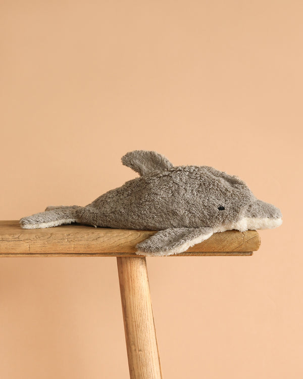 The Senger Naturwelt Cuddly Animal - Dolphin, a gray plush toy with a white belly made from sustainable materials, lies on its side on a wooden surface against a plain beige background.