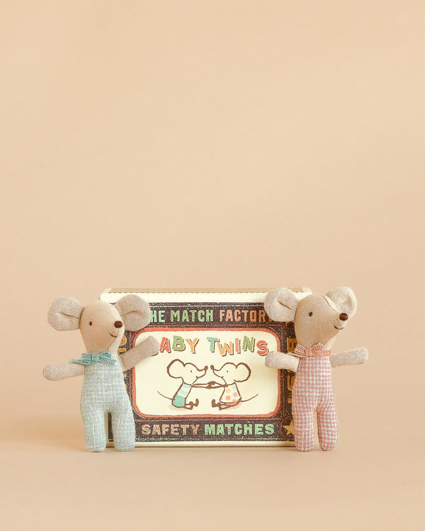 Two small Maileg Twins, Baby Mice in Matchbox - Checker—one in blue and one in pink—stand on either side of a vintage-style matchbox bed labeled “Baby Twins Safety Matches” with an illustration of two mice. The background is a warm beige.