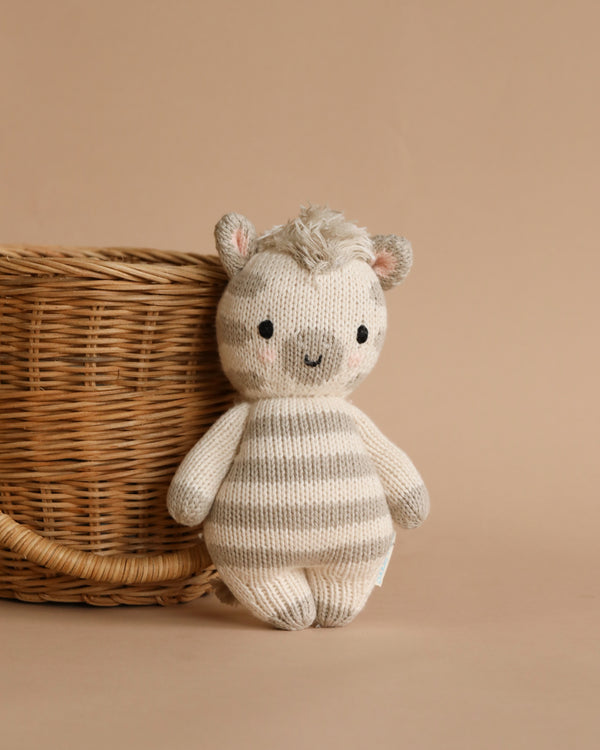 A hand-knit plush toy resembling a Cuddle + Kind Baby Zebra with striped patterns, standing next to a woven basket on a beige background.