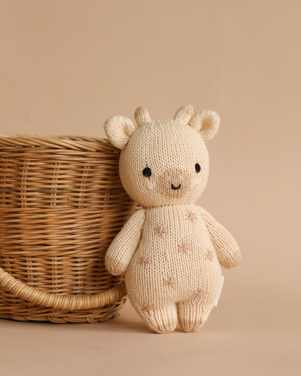 A hand-knit Cuddle + Kind Baby Giraffe, crafted from Peruvian cotton yarn, with a smiling face and button eyes, stands beside a wicker basket on a beige background.
