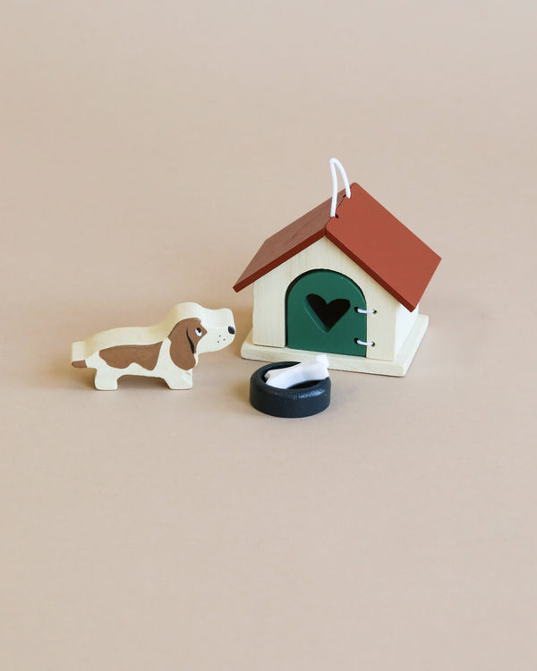 A wooden toy basset hound dog next to a small Doghouse Set with an opening door, featuring a heart-shaped cutout and a red roof on a light beige background. A black round object