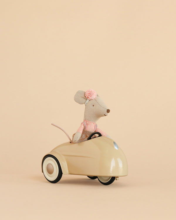 A Maileg Little Sister Driver Set, featuring a small mouse in a pink dress with a flower headband, is displayed sitting in a cream-colored car against a plain beige background.