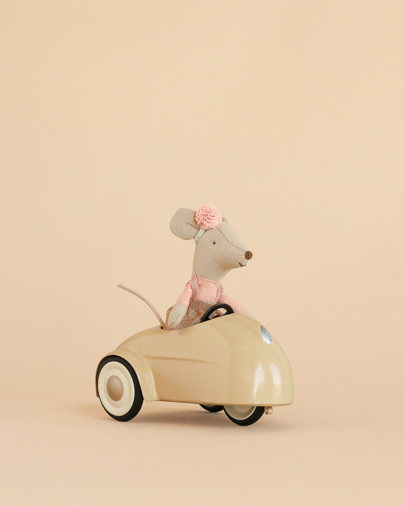 A Maileg Little Sister Driver Set, featuring a small mouse in a pink dress with a flower headband, is displayed sitting in a cream-colored car against a plain beige background.