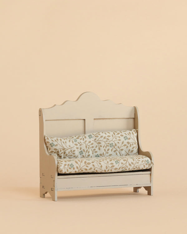 The Maileg Kitchen Bench, Mouse - Light Sand is a small wooden bench featuring a decorative backrest and soft, floral-patterned cushions, set against a plain beige background to create an inviting and cosy atmosphere.