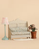 The Maileg Cozy Corner Set features a small sofa with a floral cushion, a Maileg Vintage Floor Lamp with a pink patterned shade, a potted plant, and a Maileg Lace Rug, all arranged gracefully on a beige background.