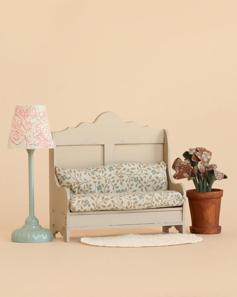 The Maileg Cozy Corner Set features a small sofa with a floral cushion, a Maileg Vintage Floor Lamp with a pink patterned shade, a potted plant, and a Maileg Lace Rug, all arranged gracefully on a beige background.
