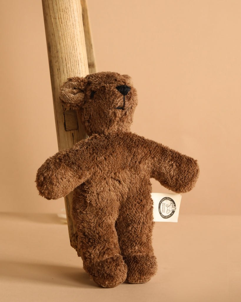 The Senger Naturwelt Stuffed Animal - Brown Bear, made from eco-friendly quality materials, features a white tag on its arm and leans against a wooden plank with a beige background.