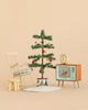 The Maileg Cozy Christmas Corner, featuring a small Christmas tree with ornaments nestled between a wooden rocking chair and a vintage TV topped with an old-fashioned alarm clock—all arranged on a round white rug against a beige backdrop—perfectly captures the charm of Maileg dollhouse accessories.