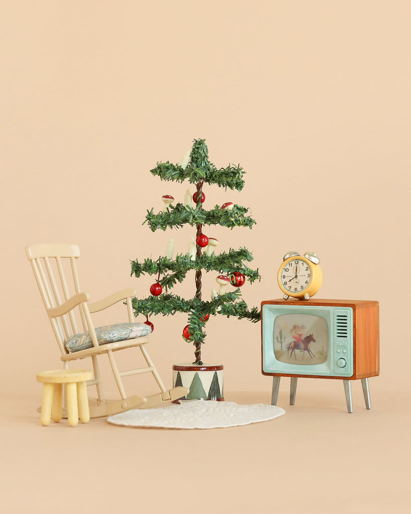 The Maileg Cozy Christmas Corner, featuring a small Christmas tree with ornaments nestled between a wooden rocking chair and a vintage TV topped with an old-fashioned alarm clock—all arranged on a round white rug against a beige backdrop—perfectly captures the charm of Maileg dollhouse accessories.