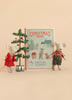 Two perfectly Mouse Size toy mice stand side by side next to a charming Maileg Christmas Tree and a copy of “Christmas Tales Bedtime Stories.” One is dressed in red overalls, while the other wears gray. Set against a warm beige background, this cozy scene showcases the delightful Maileg Christmas Twins Set.