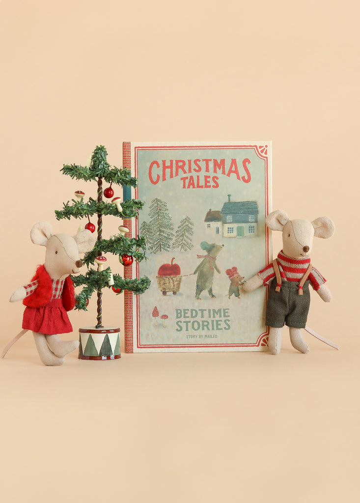 Two perfectly Mouse Size toy mice stand side by side next to a charming Maileg Christmas Tree and a copy of “Christmas Tales Bedtime Stories.” One is dressed in red overalls, while the other wears gray. Set against a warm beige background, this cozy scene showcases the delightful Maileg Christmas Twins Set.