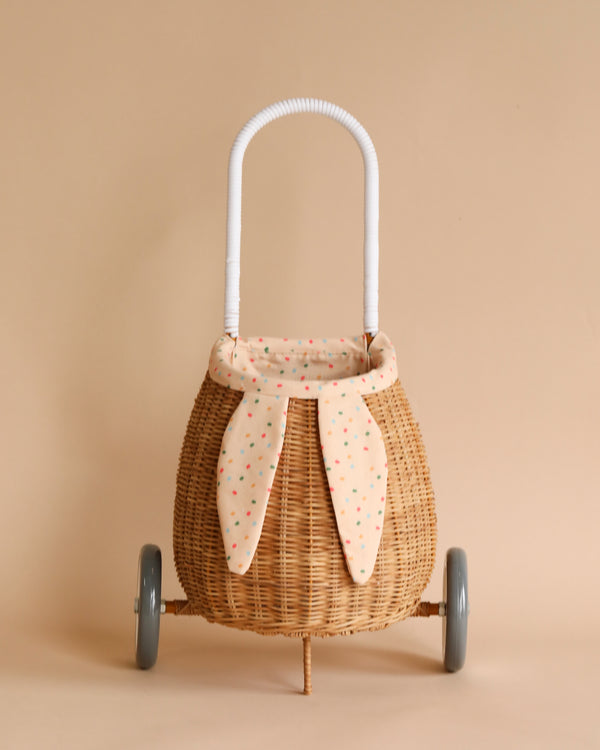 A quaint Rattan Bunny Luggy With Lining – Gumdrop, with a white wrapped handle and adorned with a polka-dotted fabric bow, set against a light tan background.