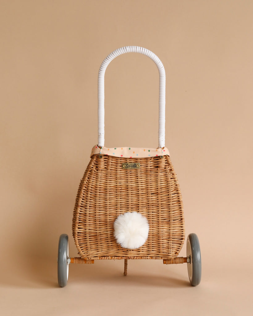 A vintage-style Rattan Bunny Luggy With Lining – Gumdrop with a white handle and wheels, featuring a fluffy white pompom and a polka dot interior, set against a neutral tan background. Crafted from natural rattan.