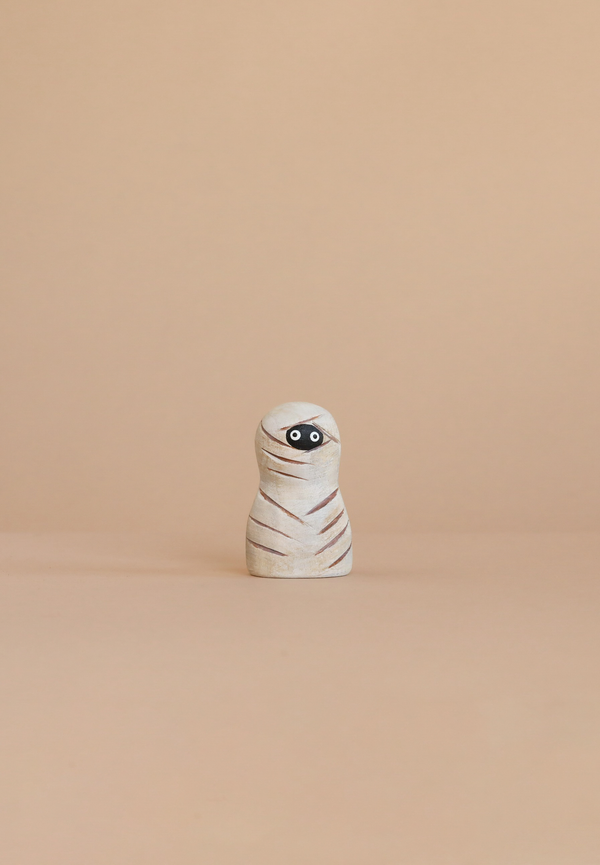 The Bumbu Handmade Wooden Enchanted Mummy, with painted white wrappings and big eyes, stands on a beige surface—perfect for imaginative play against a matching backdrop.