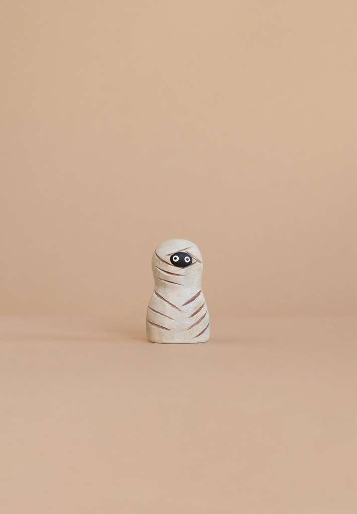 The Bumbu Handmade Wooden Enchanted Mummy, with painted white wrappings and big eyes, stands on a beige surface—perfect for imaginative play against a matching backdrop.