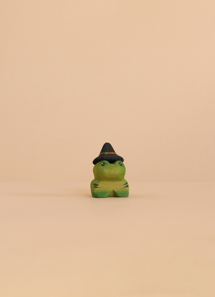 A small green Bumbu Handmade Wooden Spell Frog with a black witch hat sits on a beige surface, ideal for Waldorf or Montessori play.