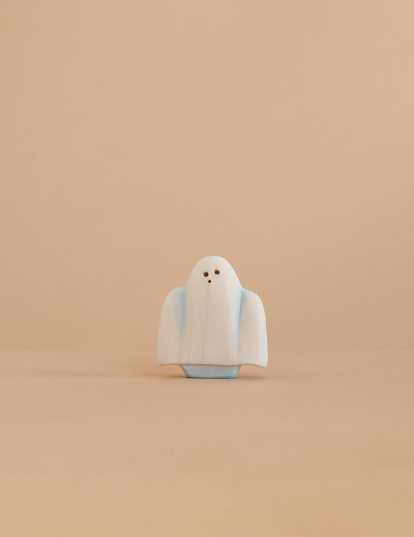 A small Bumbu Handmade Wooden Ghost with tiny black eyes stands on a beige surface against a matching plain beige background.