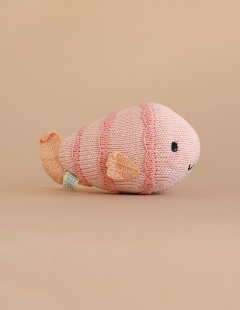 The Cuddle + Kind Baby Fish is a hand-knit pink and light peach plush with black stitched eyes and a small smile—an heirloom-quality collectible stuffed animal.