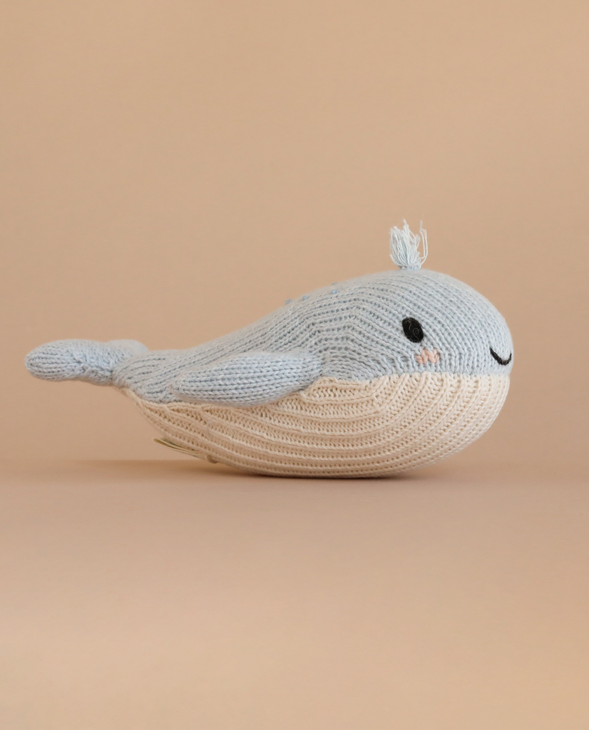 The Cuddle + Kind Baby Whale is a light blue and cream knitted plush toy with a small tail and smiling face, handmade in Peru, shown on a plain beige background.