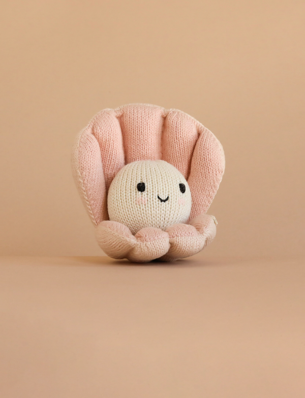 The Cuddle + Kind Baby Pearl is a hand-knit baby animal toy shaped like a smiling pearl in a pink clam shell, made from natural cotton yarn and shown on a plain beige background.