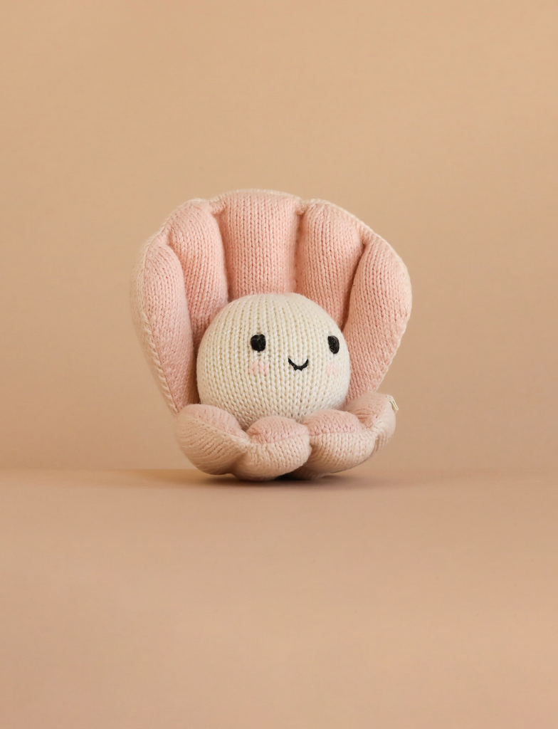 The Cuddle + Kind Baby Pearl is a hand-knit baby animal toy shaped like a smiling pearl in a pink clam shell, made from natural cotton yarn and shown on a plain beige background.