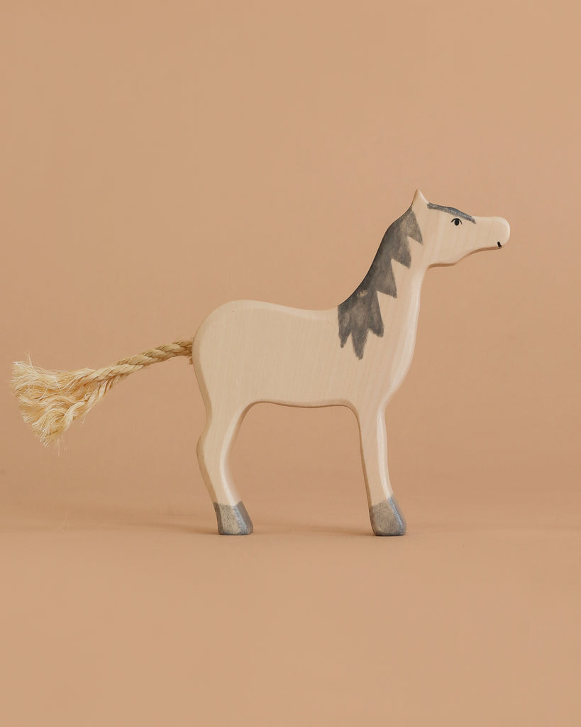A Holztiger Handmade Wooden Farm Animals Set piece—a toy horse made from maple and beech, featuring a rope tail and hand-painted gray hooves, mane, and facial details—stands on a beige surface against a beige background.