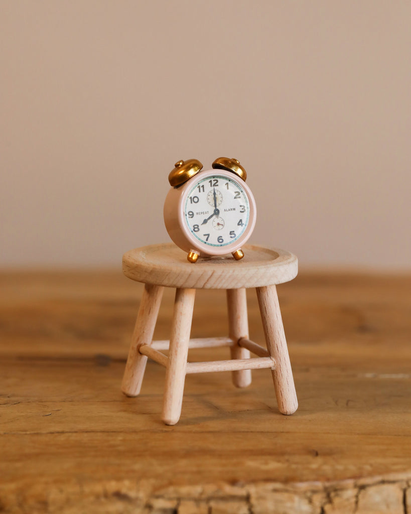 The Maileg Little Sister Bathroom Set features a small vintage-style alarm clock with gold bells on a miniature wooden stool, ideal for Maileg dollhouse furniture, displayed on a wooden surface against a plain beige background.