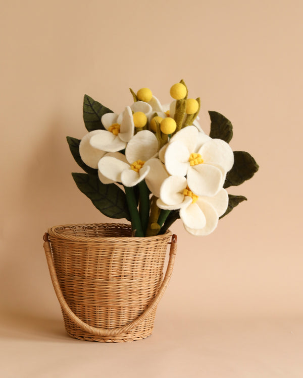 A felt flower bouquet of handmade artificial white and yellow flowers in a woven basket, placed against a neutral beige background.