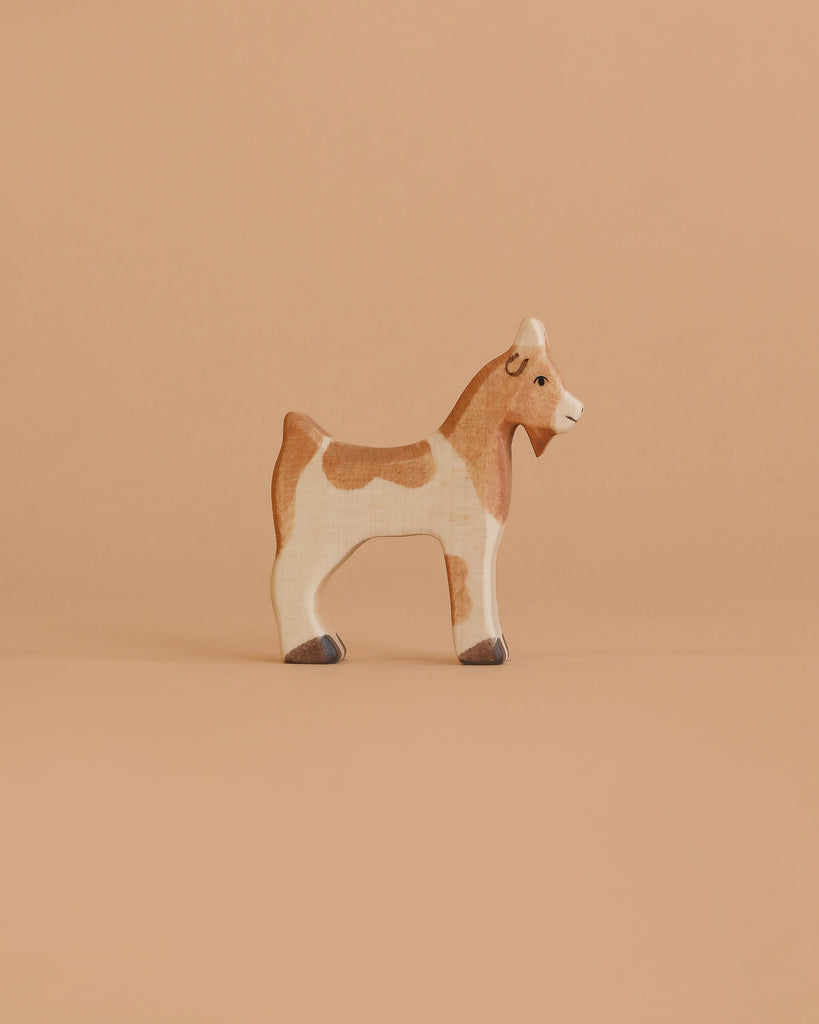 A small goat figurine from the Holztiger Handmade Wooden Farm Animals Set, crafted from maple and beech wood, stands on a beige background with light brown and white patches, simple features, and dark hooves.