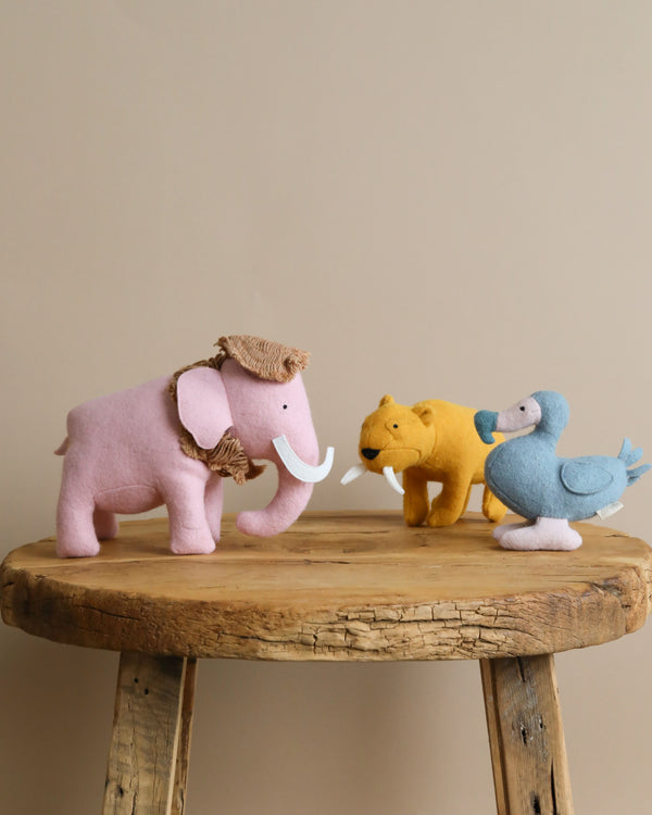 Three Olli Ella Holdie Extinct Animal toys on a wooden stool: a pink mammoth with white tusks, a yellow lion, and a blue bird, all facing each other as if in conversation.
