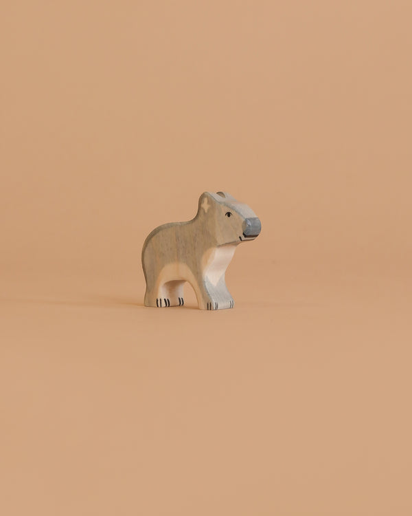 A small, carved wooden figurine of a koala stands on a plain, light peach background. Handcrafted in Europe, this Holztiger Koala, Standing features neutral gray and beige tones with simple black details.