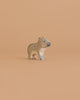A small, carved wooden figurine of a koala stands on a plain, light peach background. Handcrafted in Europe, this Holztiger Koala, Standing features neutral gray and beige tones with simple black details.