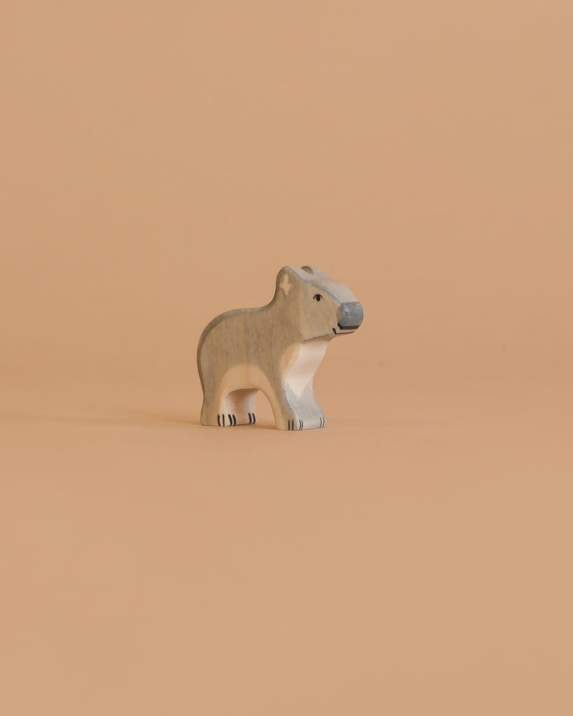 A small, carved wooden figurine of a koala stands on a plain, light peach background. Handcrafted in Europe, this Holztiger Koala, Standing features neutral gray and beige tones with simple black details.