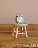 Against a neutral backdrop, the Maileg Alarm Clock For Dollhouse in mint, featuring shining metal bells and showing 10:10, rests on a rustic wooden stool. Together, they form a simple composition that highlights beauty in small details.