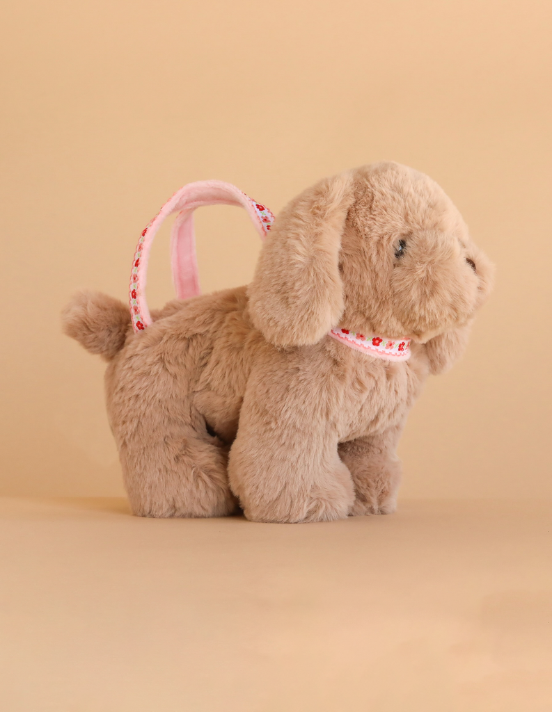 The Konges Sloejd Puppy Bag is a plush toy dog with floppy ears and a pink heart-printed strap, pictured on a neutral beige background.