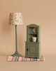 A Maileg Corner Cabinet, Mouse in light green with a vintage finish holds a small cup and plate and stands on a colorful striped rug. To its left, there is a green floor lamp with a floral-patterned lampshade. The backdrop is neutral beige, adding to the charming home decor vibe.